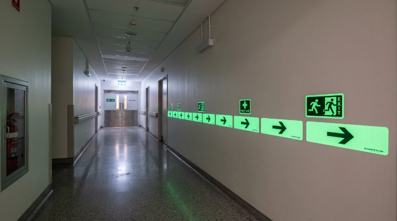 Photorealistic modern emergency corridor featuring professionally installed photoluminescent glow stickers with directional arrows and safety symbols