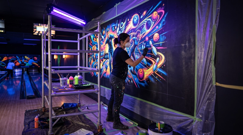 Professional commercial interior decorator applying UV Reactive Paint to a large bowling centre mural, prepared dark wall surface